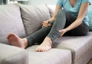 A woman sitting on her couch and holding her calf with one hand as if in pain from varicose veins