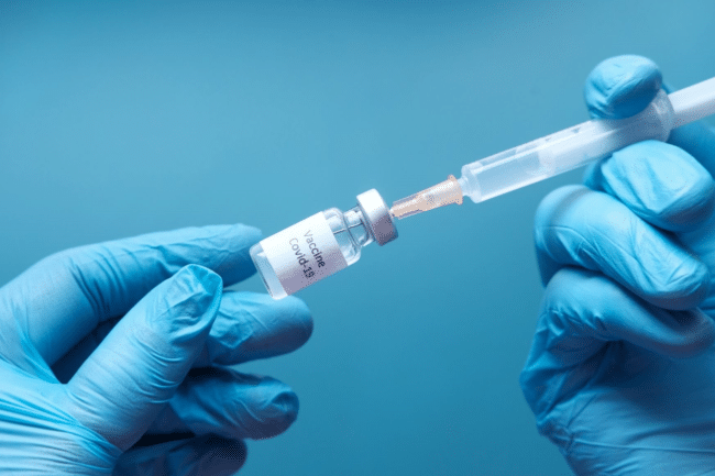 Close up of gloved hands holding a syringe and a small vial labelled COVID-19 vaccine.