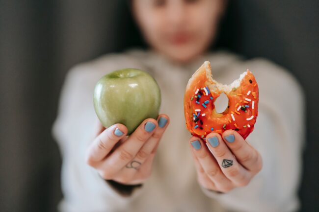 Woman holding up apple in one hand and doughnut in the other, as dietary change can be an effective varicose vein remedy