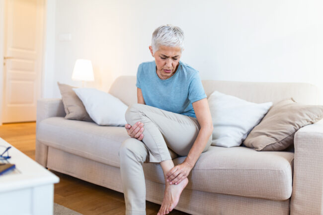 Older woman sitting on her couch, rubbing her leg in pain, wondering is it worth treating varicose veins