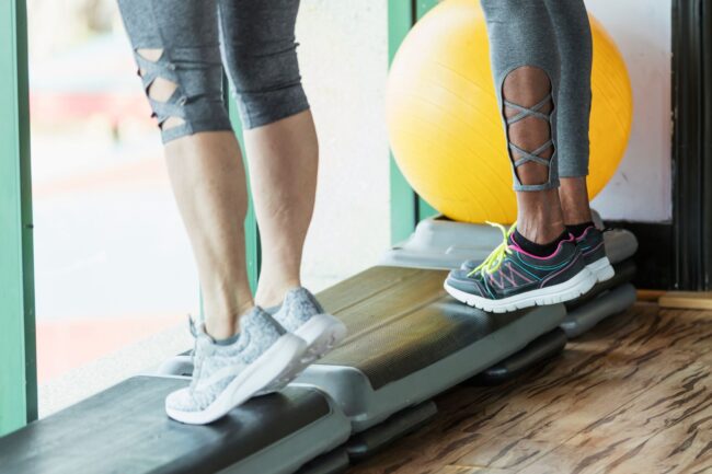 Two people standing side by side at the gym lifting their heels of the floor. Calf raises is one of the best exercises for varicose veins as it helps actively pump blood from the legs to the heart